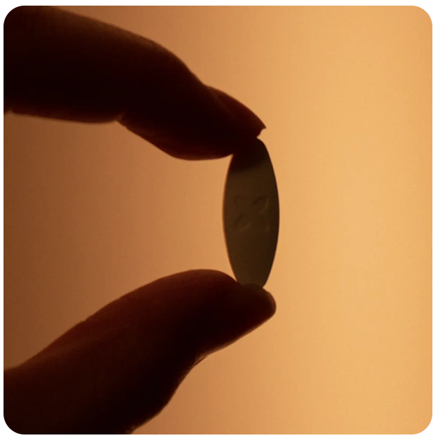 A close up silhouette of a finger and thumb holding a Boquet pill against an orange background.