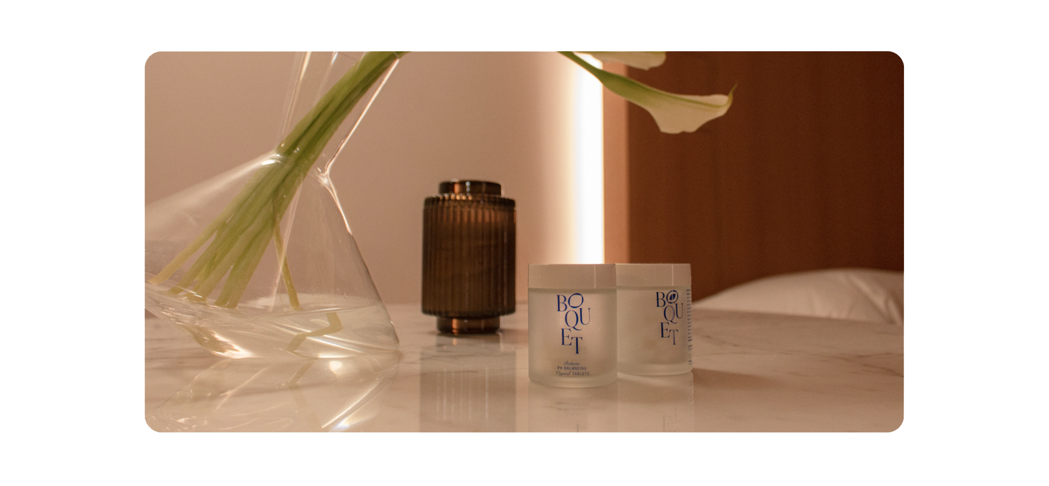 A beautiful bedroom with two frosted white glass bottles with blue Boquet logo text and a clear glass flower vase holding lilies.